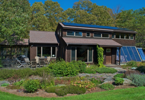 A modern two-story house with solar panels, surrounded by a lush garden and trees under a clear blue sky.