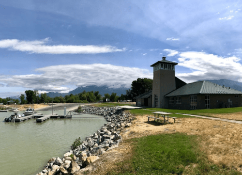 A lakeside building with a tower, surrounded by rocky shore and green trees, under a cloudy sky with mountains in the background.