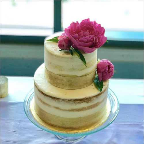 A two-tiered cake with a smooth finish, adorned with pink peonies and gold accents, displayed on a glass stand.