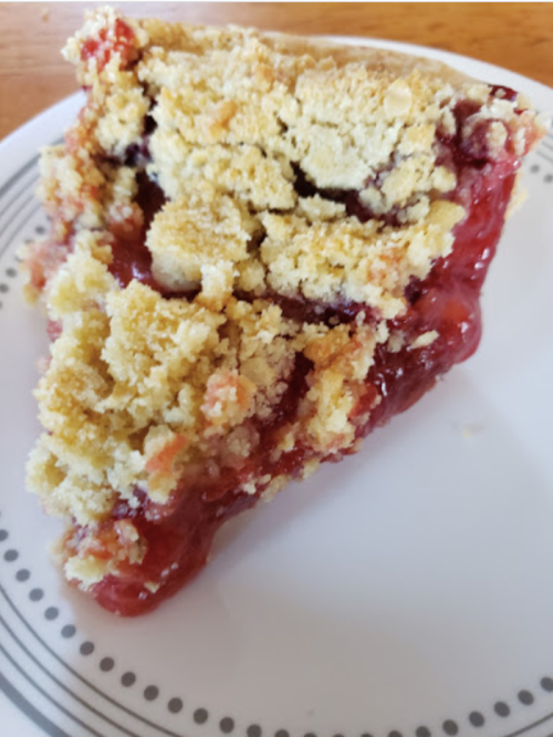 A slice of crumbly fruit pie with a red filling on a white plate with a decorative rim.