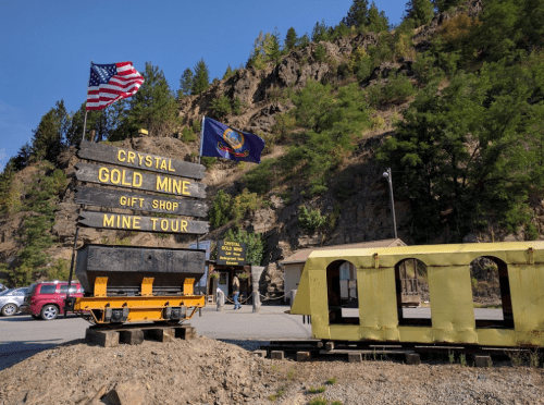 Sign for Crystal Gold Mine with American flag, gift shop, and mine tour entrance, set against a rocky hillside.