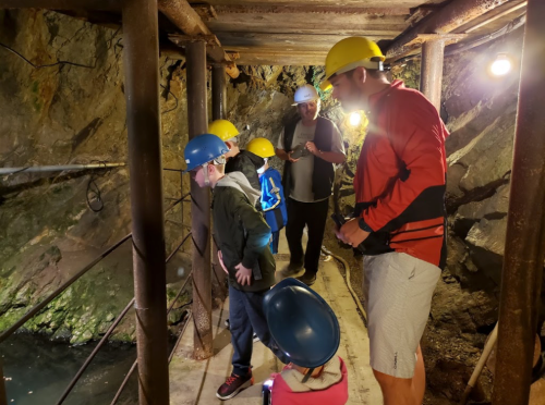 A group of people wearing helmets explores a dimly lit mine, standing on a wooden walkway near water.