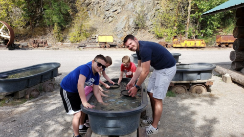 A group of children and an adult play in water-filled troughs outdoors, surrounded by trees and old mining equipment.