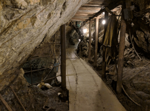 A dimly lit underground mine tunnel with wooden supports and a concrete path, surrounded by rocky walls.