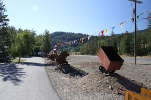 A gravel road lined with old mining carts and colorful flags, surrounded by trees and mountains in the background.