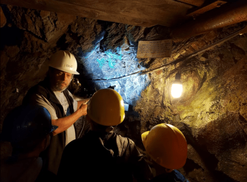 A guide explains rock formations to a group in a dimly lit mine, wearing hard hats and surrounded by rocky walls.