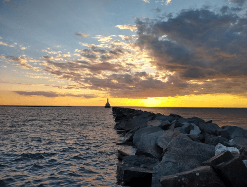A rocky jetty extends into the water, leading to a lighthouse at sunset, with colorful clouds in the sky.