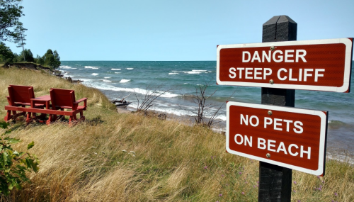 A sign warns of a steep cliff near a beach, with two red chairs overlooking the water and tall grass in the foreground.