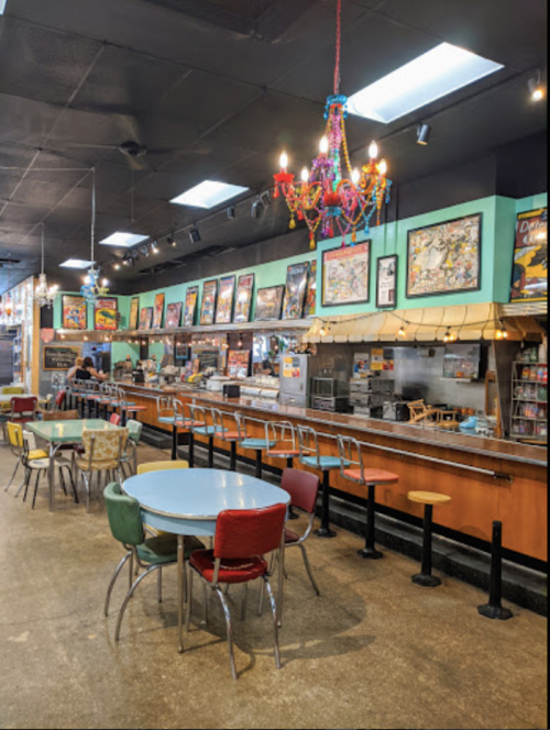 A colorful diner interior with a counter, tables, and a vibrant chandelier, adorned with framed artwork on the walls.