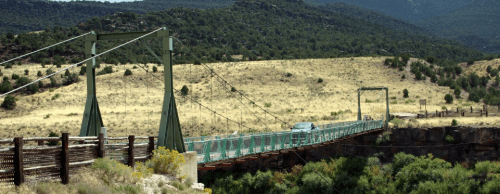 A cable-stayed bridge spans a canyon, surrounded by green hills and dry grassland under a clear sky.