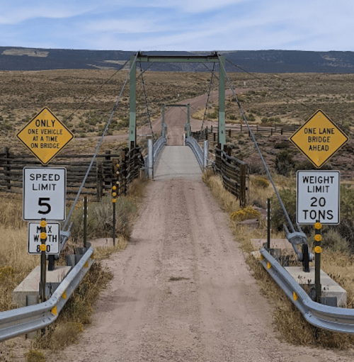 A narrow, one-lane bridge with signs for weight and speed limits, surrounded by open land and a dirt road.