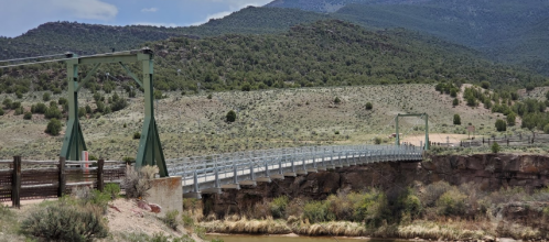 A suspension bridge spans a river, surrounded by green hills and a clear blue sky.
