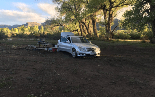 A silver car parked in a grassy area with trees and mountains in the background, alongside a small table setup.