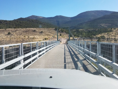 A view from a car approaching a metal bridge over a dry landscape with mountains in the background under a clear blue sky.