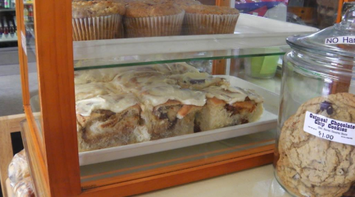 A display case with cinnamon rolls topped with icing, muffins in the background, and a jar of cookies on the side.