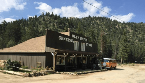 A rustic general store in Glen Haven, surrounded by trees and mountains under a blue sky. An orange vintage van is parked nearby.