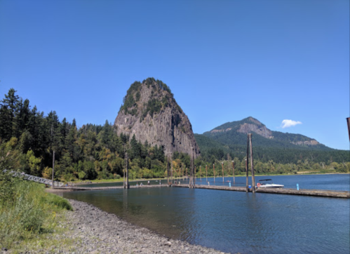 Scenic view of a river with a dock, surrounded by lush greenery and towering mountains under a clear blue sky.