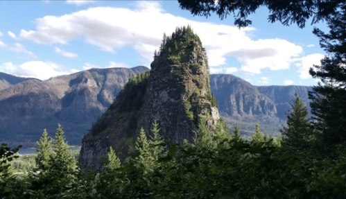 A tall, rocky peak surrounded by lush greenery and mountains under a blue sky with scattered clouds.