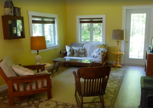 Cozy living room with yellow walls, wooden furniture, a glass coffee table, and large windows letting in natural light.