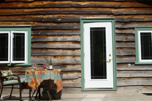 A rustic log cabin wall with a white door and two windows, featuring a table set with drinks and a colorful tablecloth.