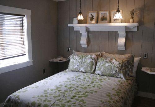 Cozy bedroom with a double bed, green patterned bedding, wooden walls, and decorative shelves with framed art.