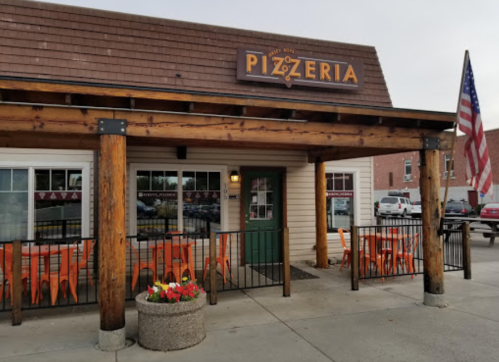Exterior of a pizzeria with a wooden facade, orange chairs outside, and an American flag.
