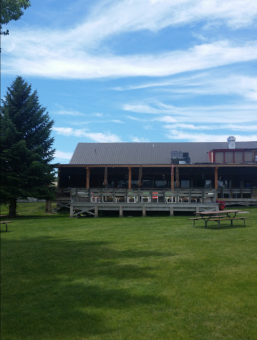 A wooden building with a deck, surrounded by green grass and trees, under a blue sky with clouds.
