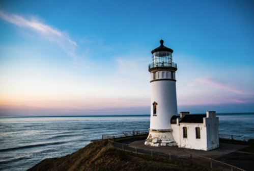 A white lighthouse stands on a cliff by the ocean, with a colorful sunset sky in the background.