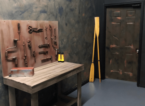A dimly lit room with a wooden table, tools on the wall, and a door beside two yellow paddles.