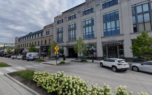 A street view of a modern building with shops, surrounded by greenery and parked cars under a cloudy sky.