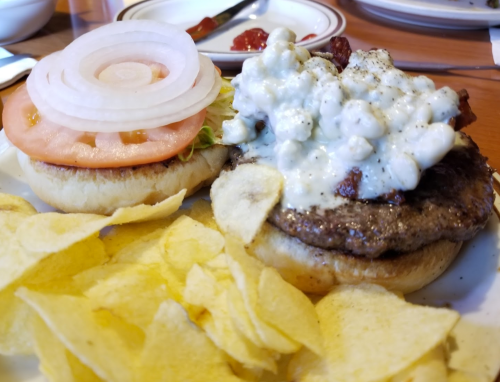 A burger with a beef patty topped with creamy sauce, onions, and tomato, served with a side of potato chips.