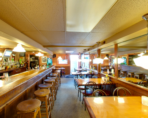 A cozy bar interior with wooden stools, tables, and a mirrored back wall, featuring soft lighting and a welcoming atmosphere.