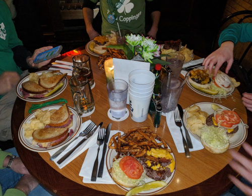 A table set with plates of burgers, sandwiches, sweet potato fries, and drinks, surrounded by hands reaching for food.