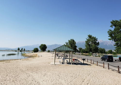A sandy beach with a gazebo, trees, and mountains in the background under a clear blue sky.