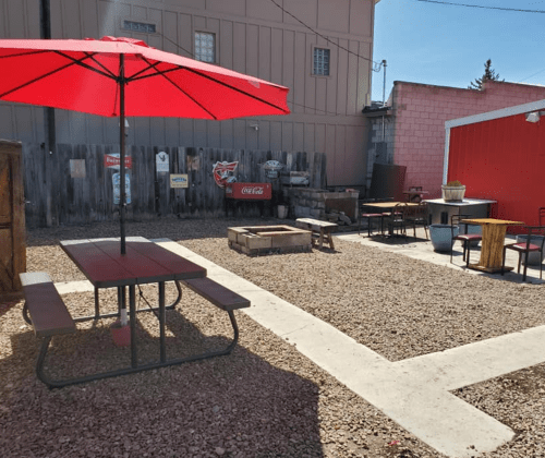 Outdoor seating area with a red umbrella, picnic tables, and a fire pit, surrounded by gravel and rustic decor.