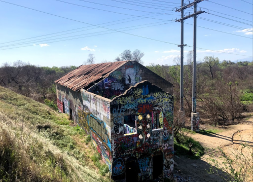 Abandoned building covered in colorful graffiti, surrounded by greenery and power lines under a clear blue sky.