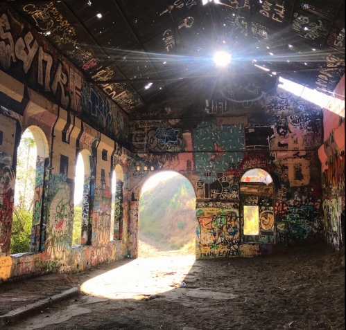 Sunlight streams through a roof with missing tiles, illuminating a graffiti-covered interior of an abandoned building.
