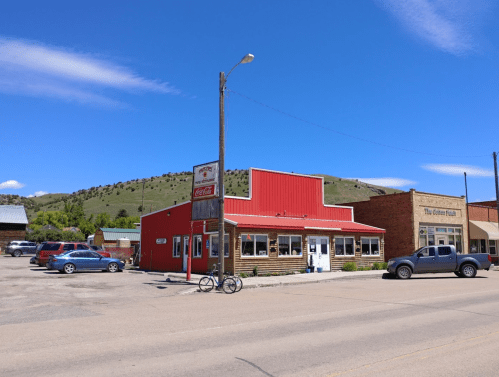 A red-roofed building in a small town, with parked cars and mountains in the background under a clear blue sky.