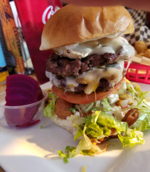 A towering burger with two patties, cheese, lettuce, tomato, and a side of pickled vegetables on a plate.