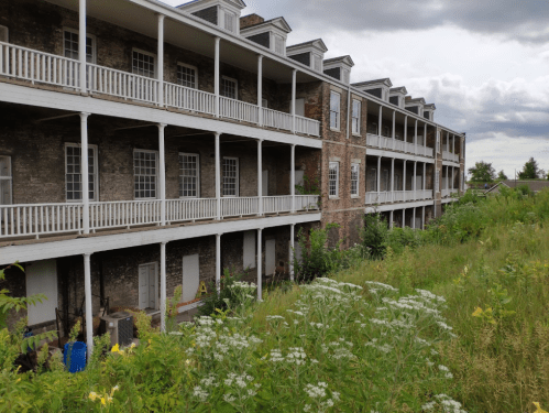 Abandoned building with multiple balconies, overgrown grass, and wildflowers in the foreground under a cloudy sky.