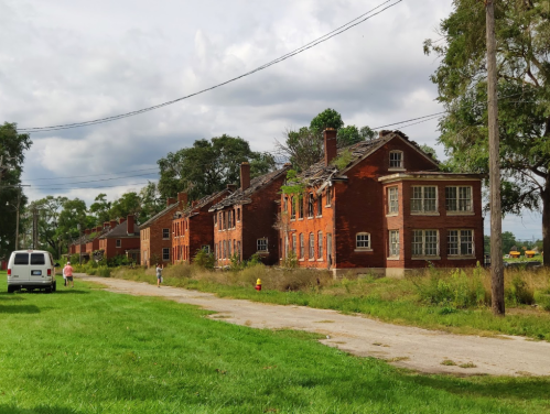 Abandoned red brick buildings with overgrown grass and trees, along a dirt road under a cloudy sky.