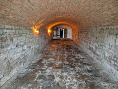 A dimly lit stone tunnel with a brick archway and wet floor, leading to a doorway at the far end.