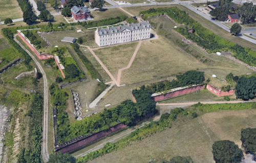 Aerial view of a historic fort with a large building, surrounded by greenery and earthworks, set in an open landscape.