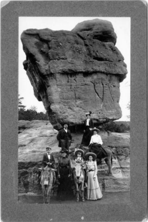 A historic black-and-white photo of six people on horseback near a large rock formation.