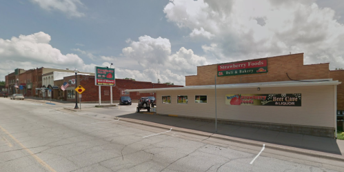 A street view of Strawberry Foods deli and bakery, with nearby shops and a cloudy sky in the background.
