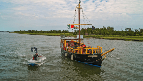 A pirate-themed ship sails on a river, with a small boat nearby and passengers enjoying the ride.