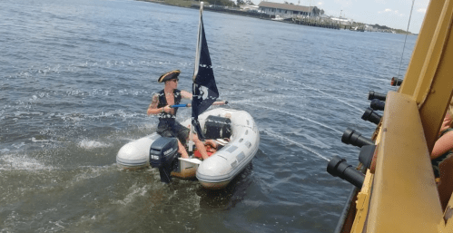 A person in a pirate hat steers a small inflatable boat with a black flag on a sunny day near the shore.