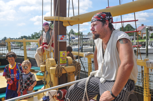 Two pirates on a ship with children dressed as pirates, enjoying a sunny day by the water.