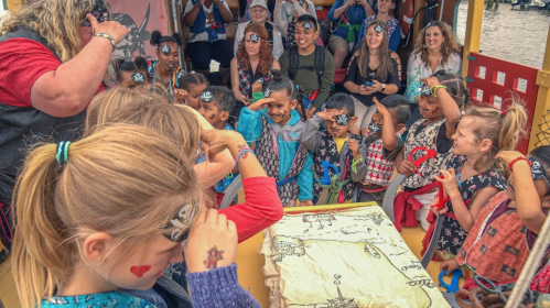 A group of children in pirate costumes on a boat, engaging with adults and looking at a treasure map.