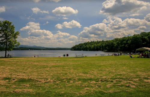 A serene lake scene with people enjoying the beach, green grass, and fluffy clouds in a blue sky.